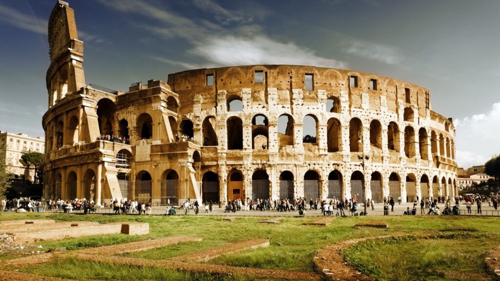 The Colosseum in Rome, Italy, stands under a partly cloudy sky, with crowds of people walking around its ancient stone structure and ruins in the grassy foreground.