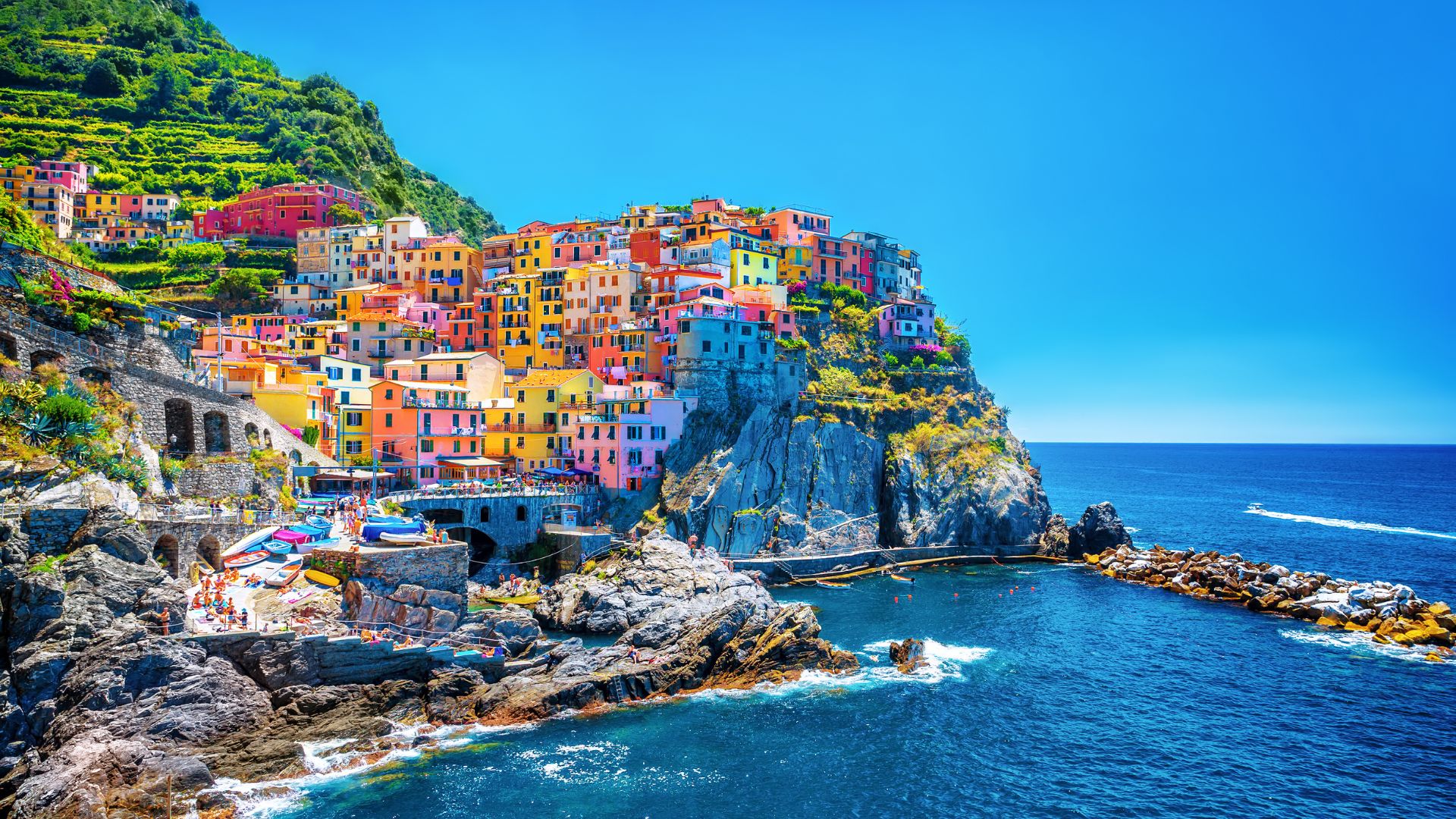Colorful houses are perched on a rocky cliff above the bright blue sea in Amalfi, with terraced green hills rising behind them under a clear, sunny sky. Boats and people near the water’s edge evoke the charm of an NCL Mediterranean cruise.